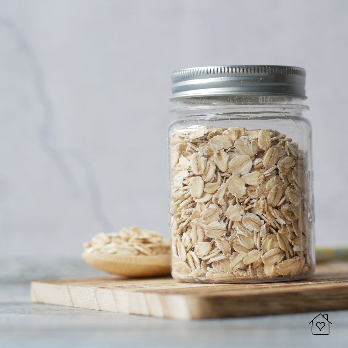 Clear glass pantry jar filled with rolled oats, sitting on a cutting board with a spoon beside it