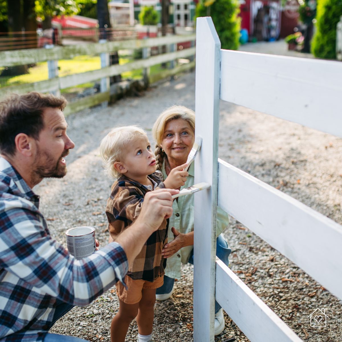 Toddler helping two adults paint a white wooden fence outside, using small brushes