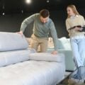 Man pressing a sofa cushion while shopping in a furniture showroom as a woman reviews a product brochure.