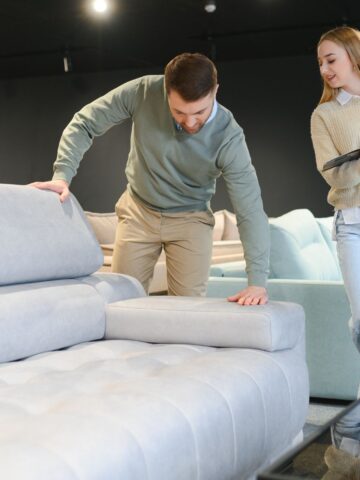 Man pressing a sofa cushion while shopping in a furniture showroom as a woman reviews a product brochure.