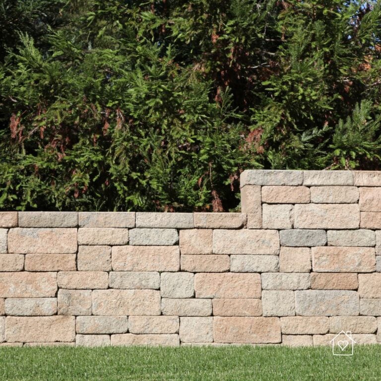 Straight tan block segmental retaining wall holding back soil with evergreen trees behind and lawn in front.