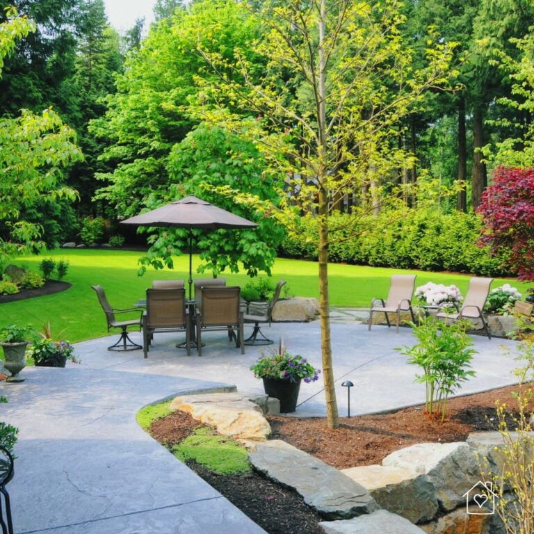 Outdoor patio with table, umbrella, lounge chairs, and garden beds opening to a wide, green lawn.