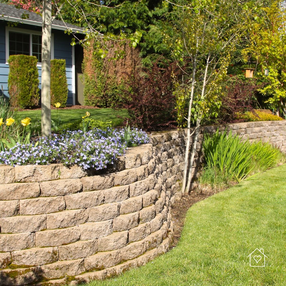Curved stacked-block retaining wall forming a raised flower bed and landscaped border along a residential lawn.