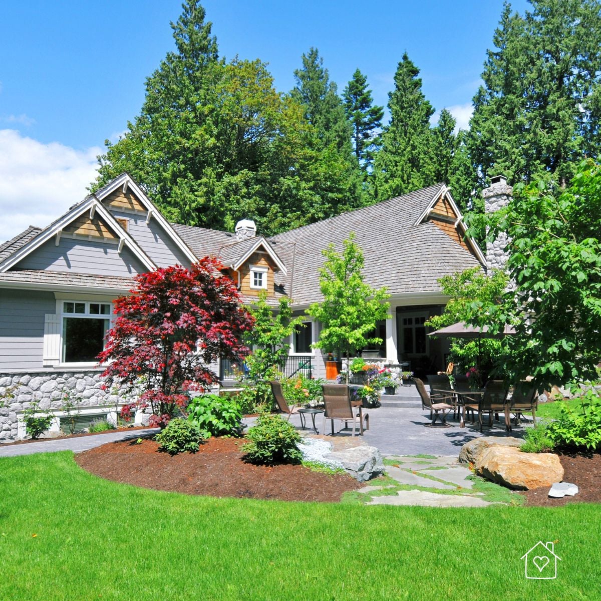 Home with a landscaped yard featuring a paved seating area, trees, shrubs, and curved garden beds.