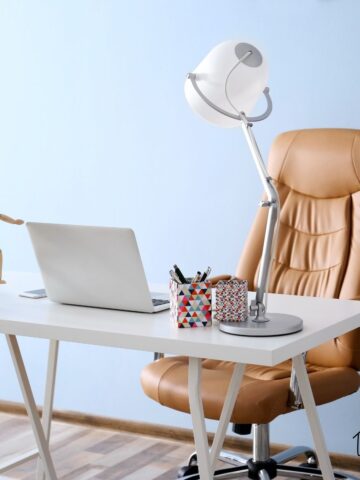 Tan leather high-back office chair beside a white minimalist desk with laptop and task lamp in a simple home office.