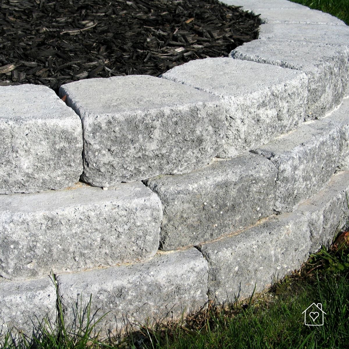 Close-up of gray concrete retaining wall blocks with textured faces and capstones beside grass and a walkway edge.