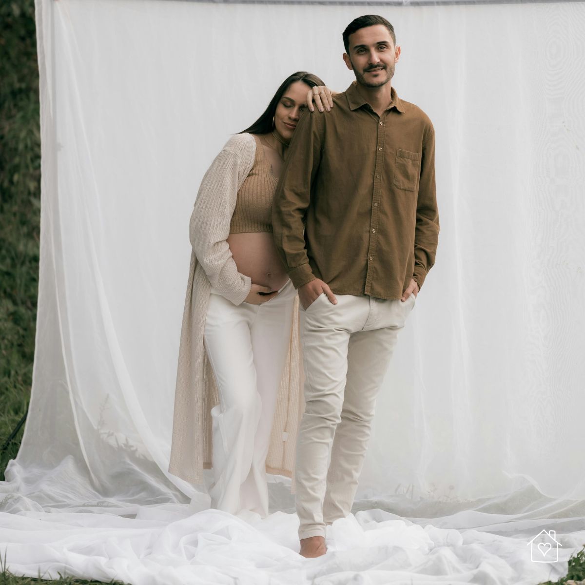 pregnant couple posing in front of a sheer fabric backdrop for an at-home maternity photoshoot