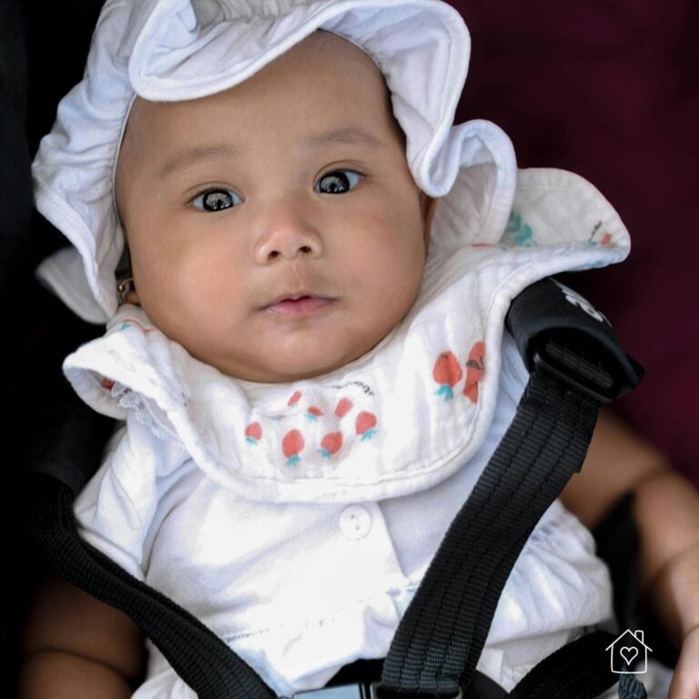 Infant wearing a white outfit and hat secured safely in a rear-facing car seat.