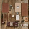 Flat lay of handmade leather accessories on rustic wood, including wallets, watch straps, notebook cover, and small everyday carry items.