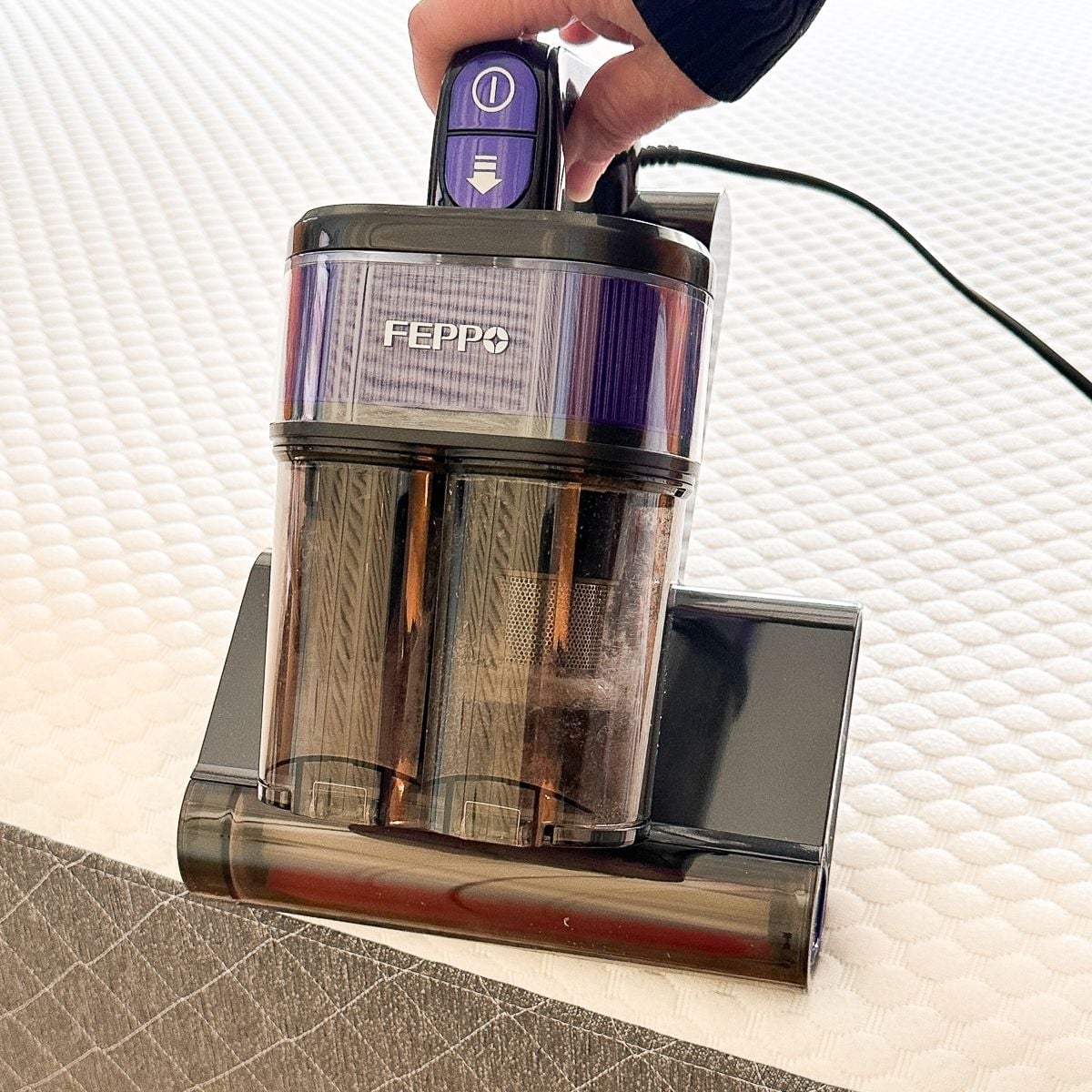 A hand holding the FEPPO mattress vacuum upright on a mattress, with the transparent dust container showing visibly dirty filter cylinders after cleaning