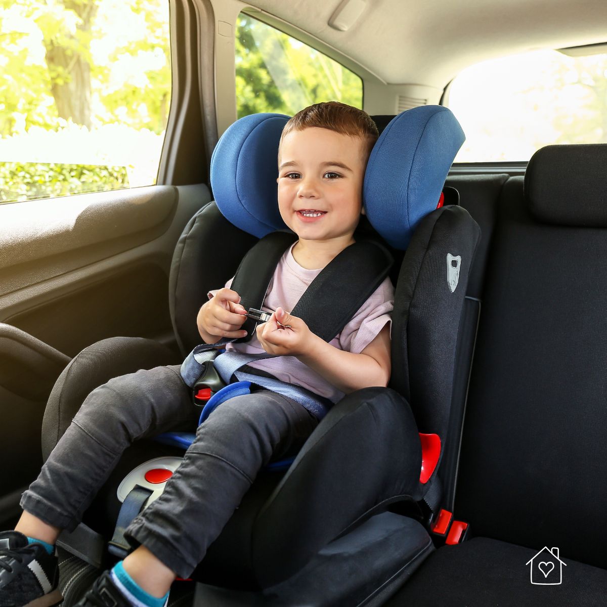 Smiling toddler seated in a booster seat with the seat belt positioned securely in the back seat of a car.