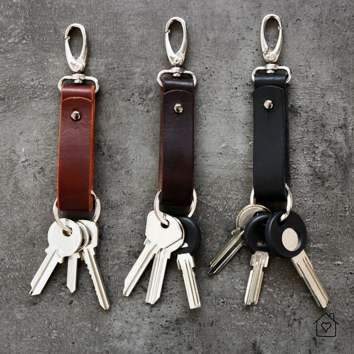 Three handmade leather keychains in brown and black with metal clips and keys on a gray textured background.