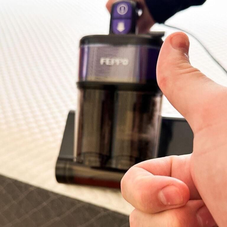 A hand giving a thumbs up in front of the FEPPO mattress vacuum cleaner sitting on a white mattress
