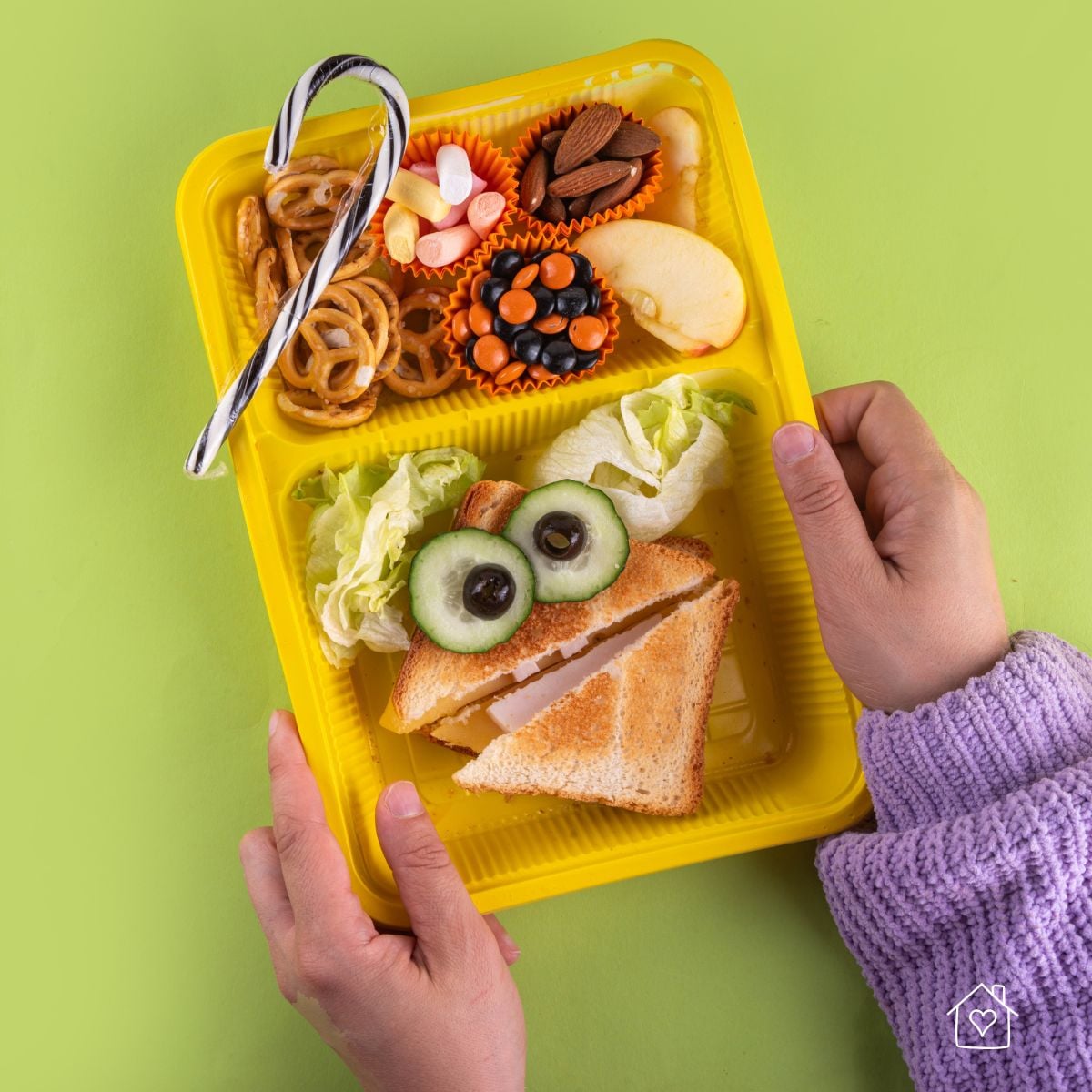Child holding a yellow school lunch tray with a fun sandwich, apple slices, pretzels, almonds, candy pieces, and lettuce.