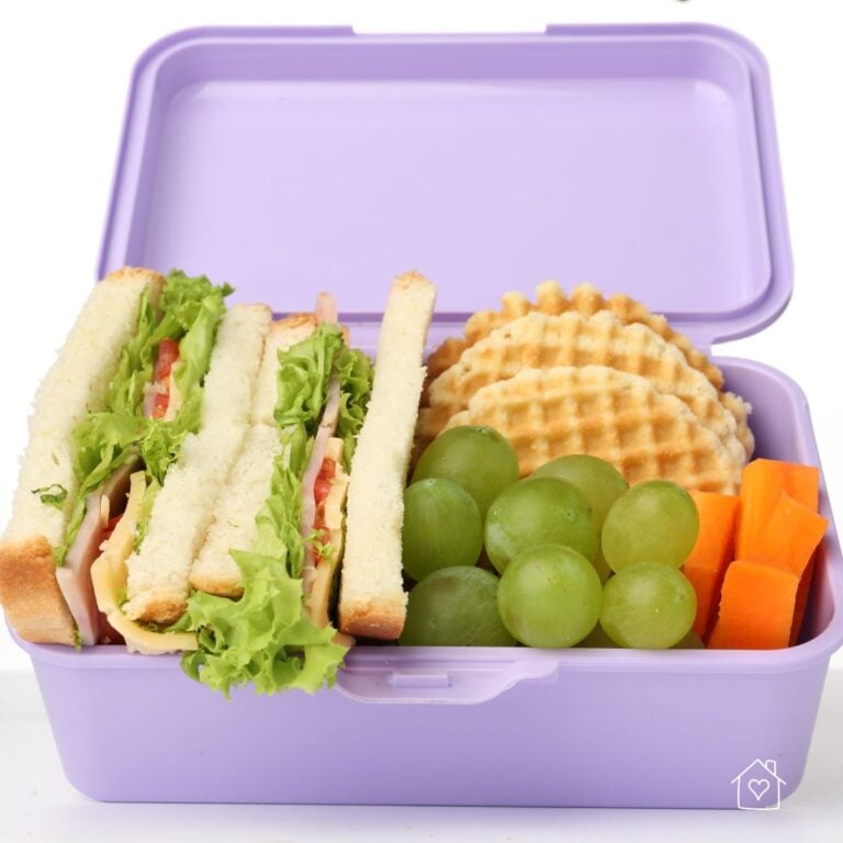 Purple lunchbox packed with sandwich slices, green grapes, waffle crackers, and carrot sticks for a school snack or lunch.