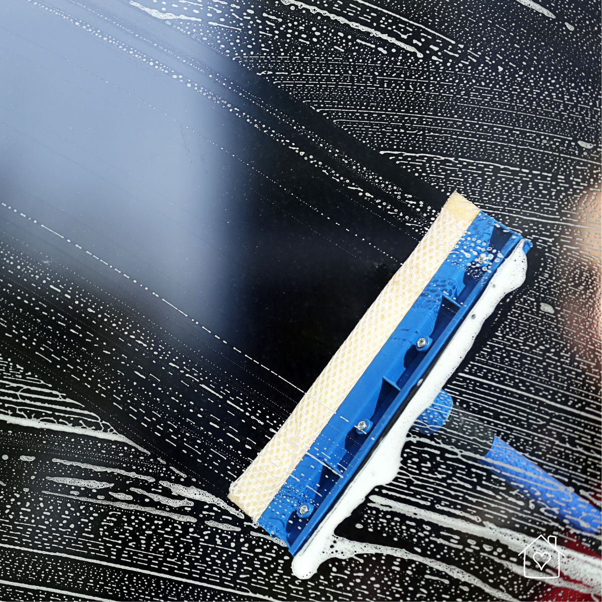 A blue and tan professional window scrubber tool cleaning a large dark exterior window covered in soapy suds