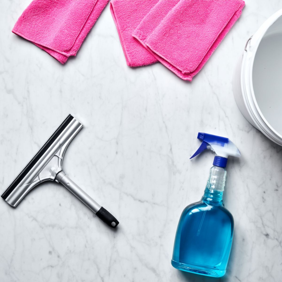 A flat lay of window cleaning supplies on a white marble surface including a silver squeegee, pink microfiber cloths, a blue spray bottle, and a white bucket
