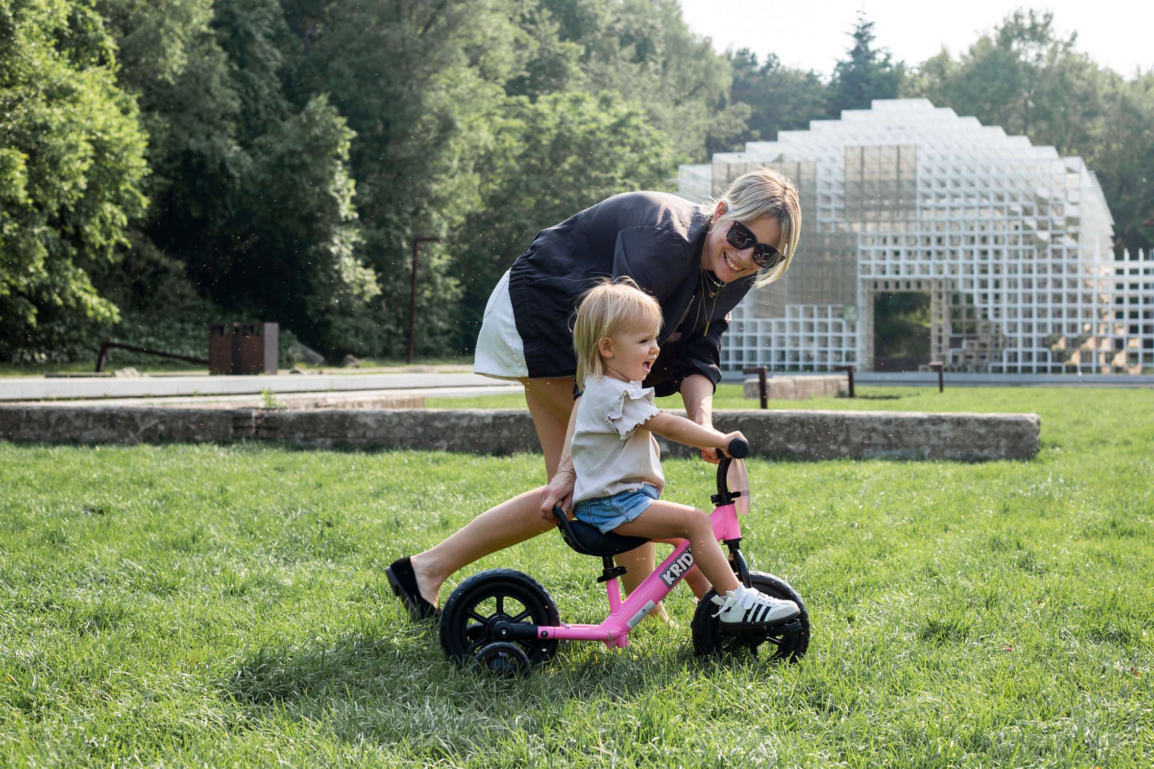 Mother helping a toddler learn to ride a pink balance bike on the grass at a park.