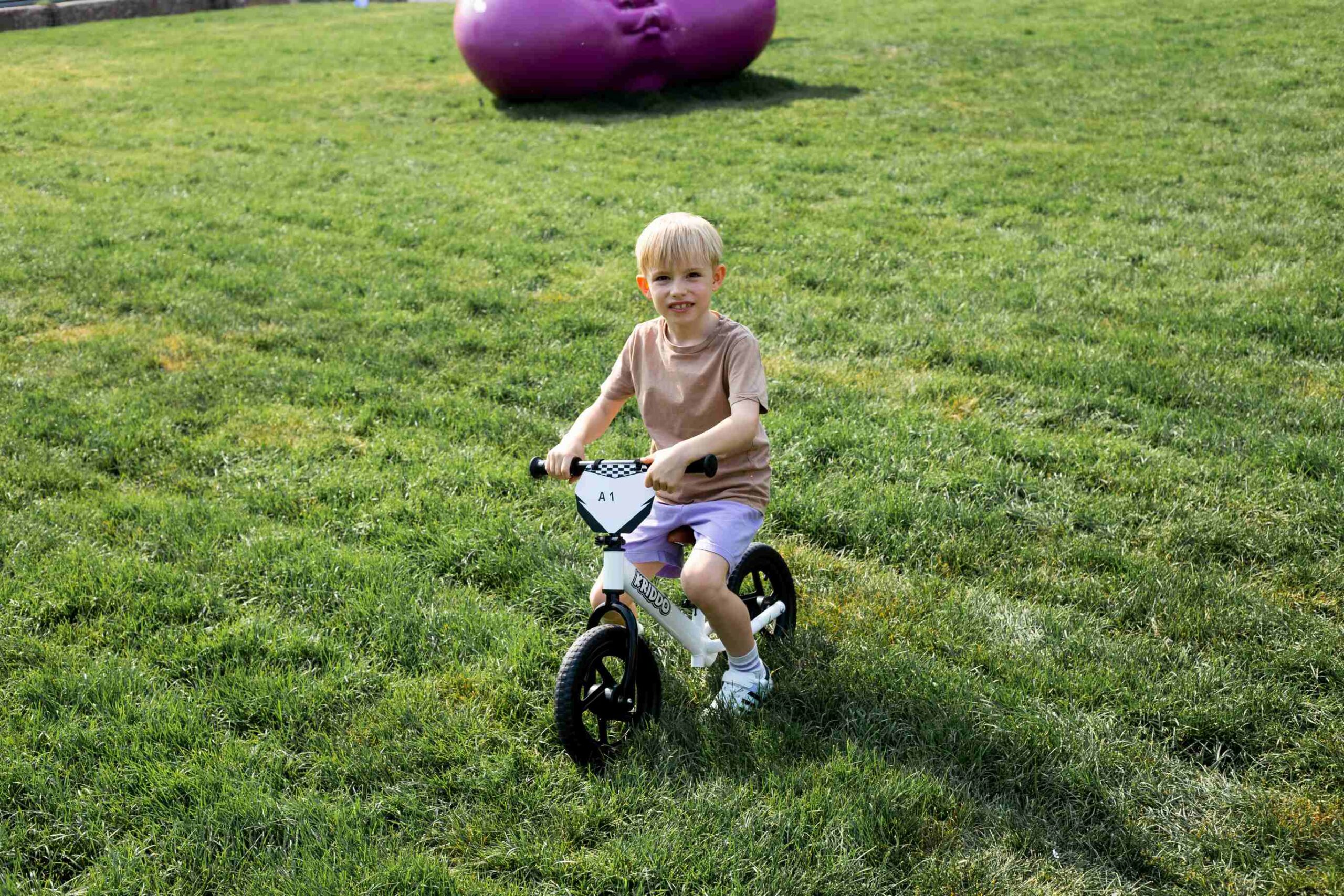 Young boy riding a white balance bike across a grassy park.