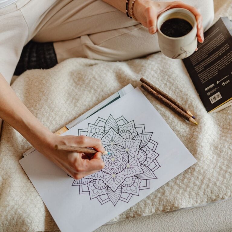 Adult coloring a mandala page with colored pencils while holding a cup of coffee in a cozy home setting