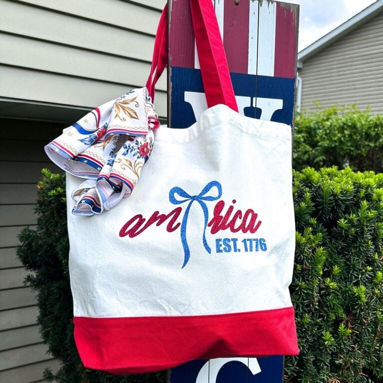 White canvas tote bag with red handles featuring glitter HTV America EST 1776 bow design hanging on patriotic blue wooden welcome sign