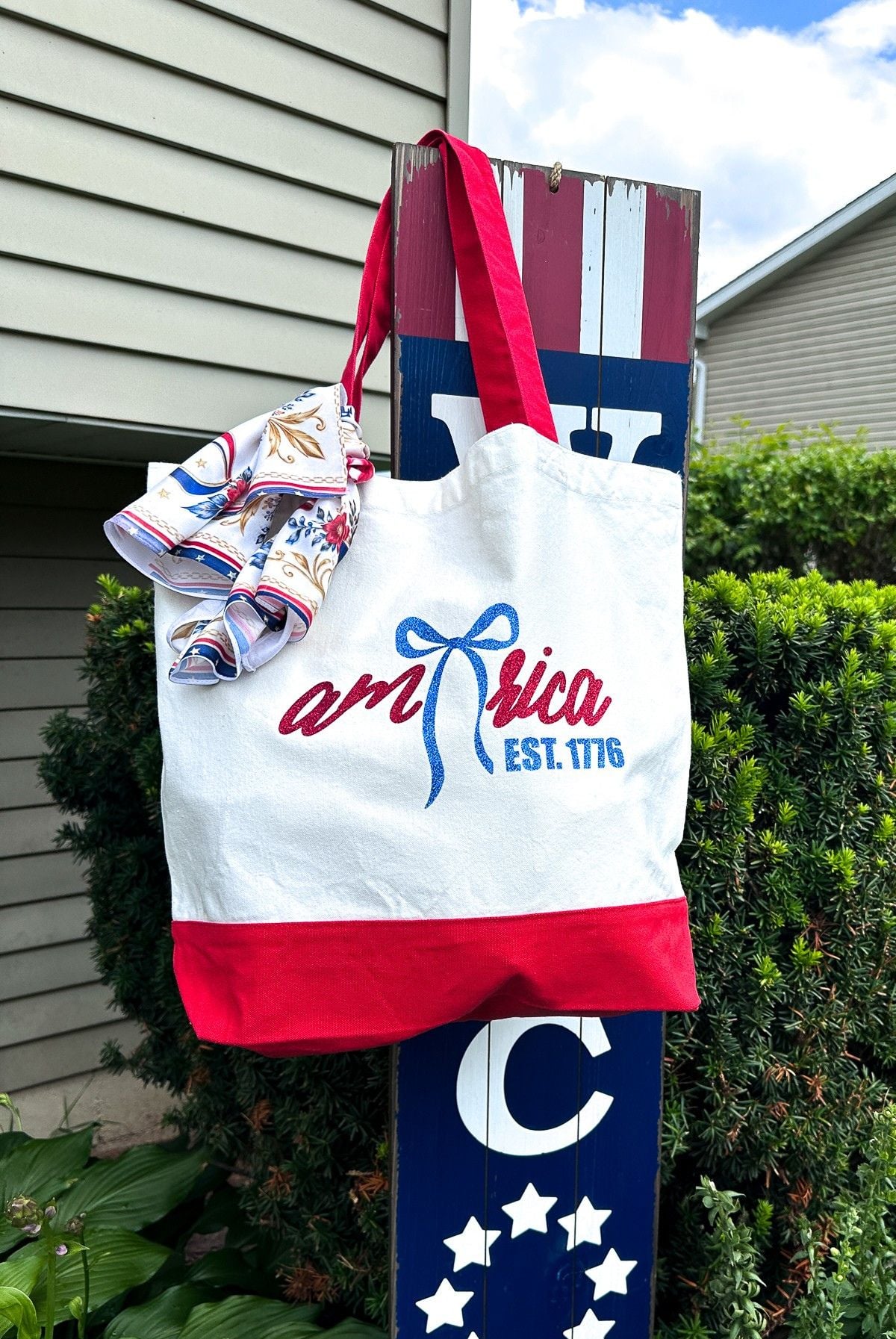 White canvas tote bag with red handles featuring glitter HTV America EST 1776 bow design hanging on patriotic blue wooden welcome sign