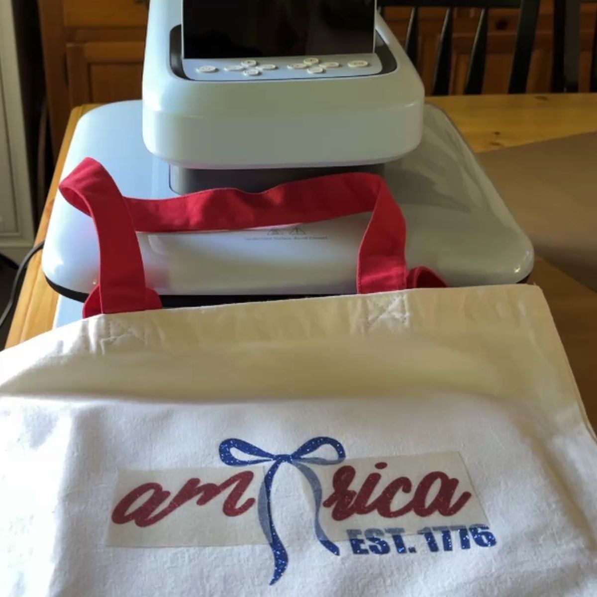 Canvas tote bag with America EST 1776 glitter HTV design positioned under HTVRont Auto Heat Press 2 with carrier sheet in place ready to press
