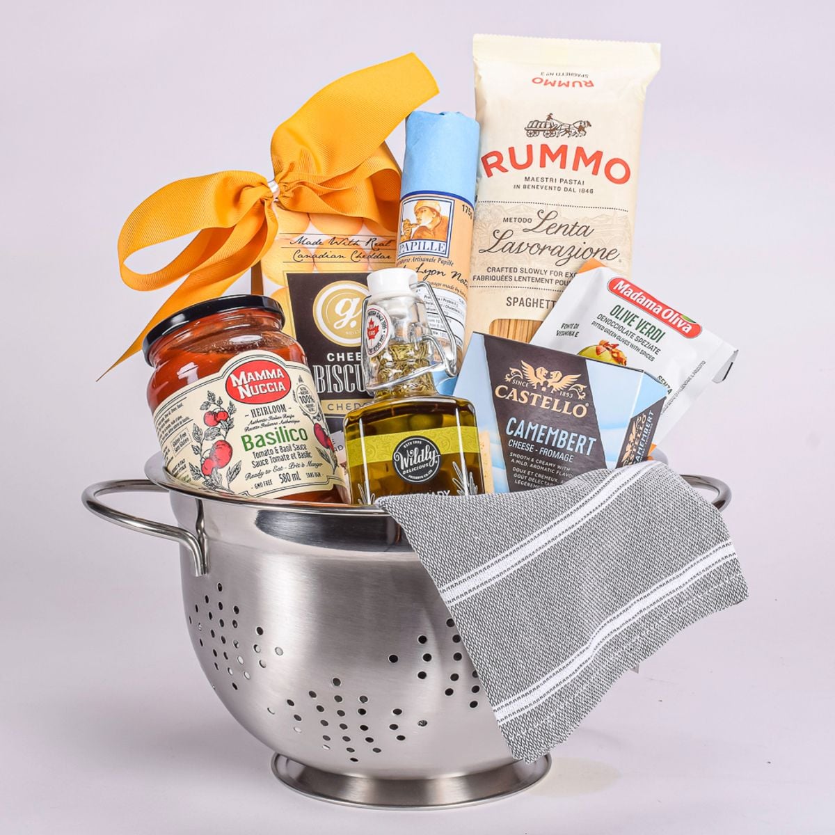 Italian housewarming gift basket with pasta, tomato basil sauce, olive oil, cheese and crackers arranged in a stainless-steel colander