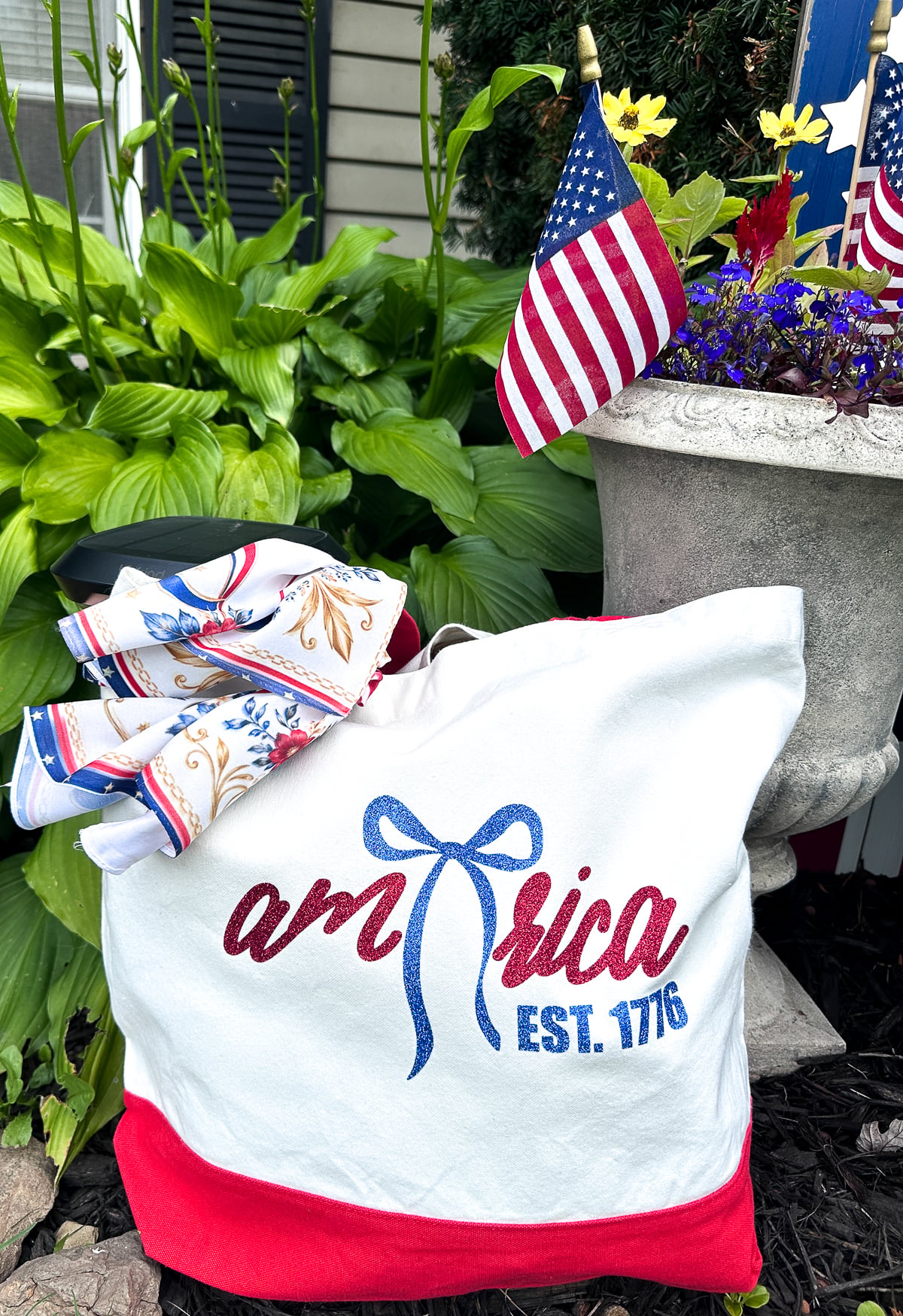 White canvas tote bag with red bottom and handles featuring glitter HTV America EST 1776 bow design displayed in garden with American flags