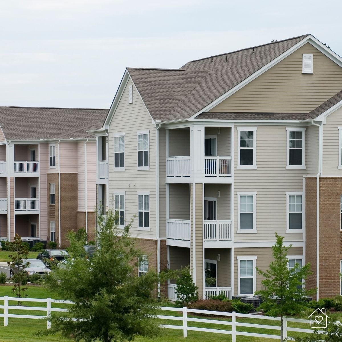 Exterior view of a multi-story apartment complex with balconies, landscaped grounds, and a white fence.