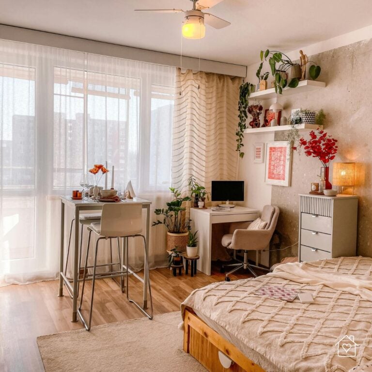 Bright studio apartment with a bed, dining table, desk, plants, shelving, and large windows.