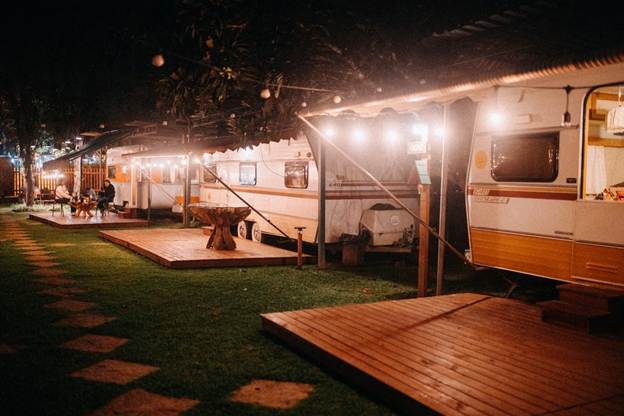 Travelers relaxing beside illuminated caravans at a campsite during an evening road trip.This image shows a nighttime caravan park with travelers gathered near a table beneath warm lights.