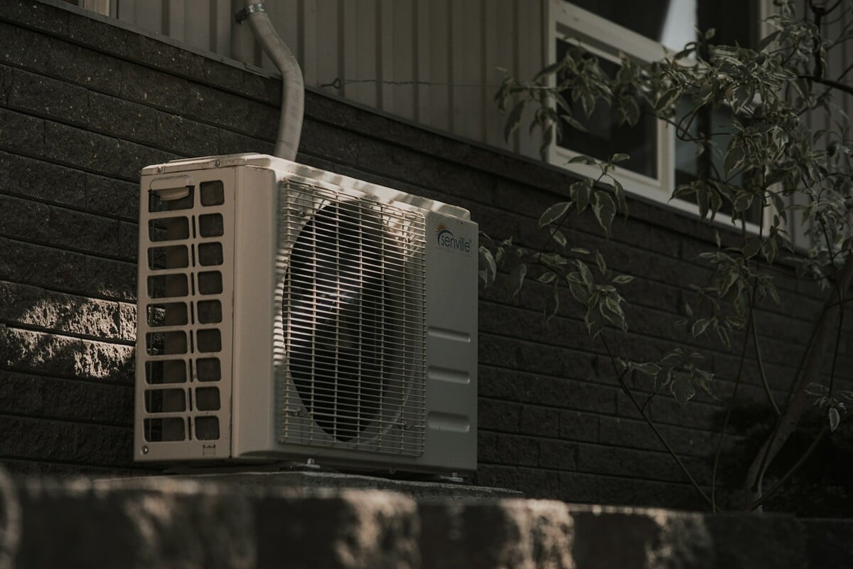 White outdoor HVAC condenser unit mounted on an exterior wall, running during a heat wave
