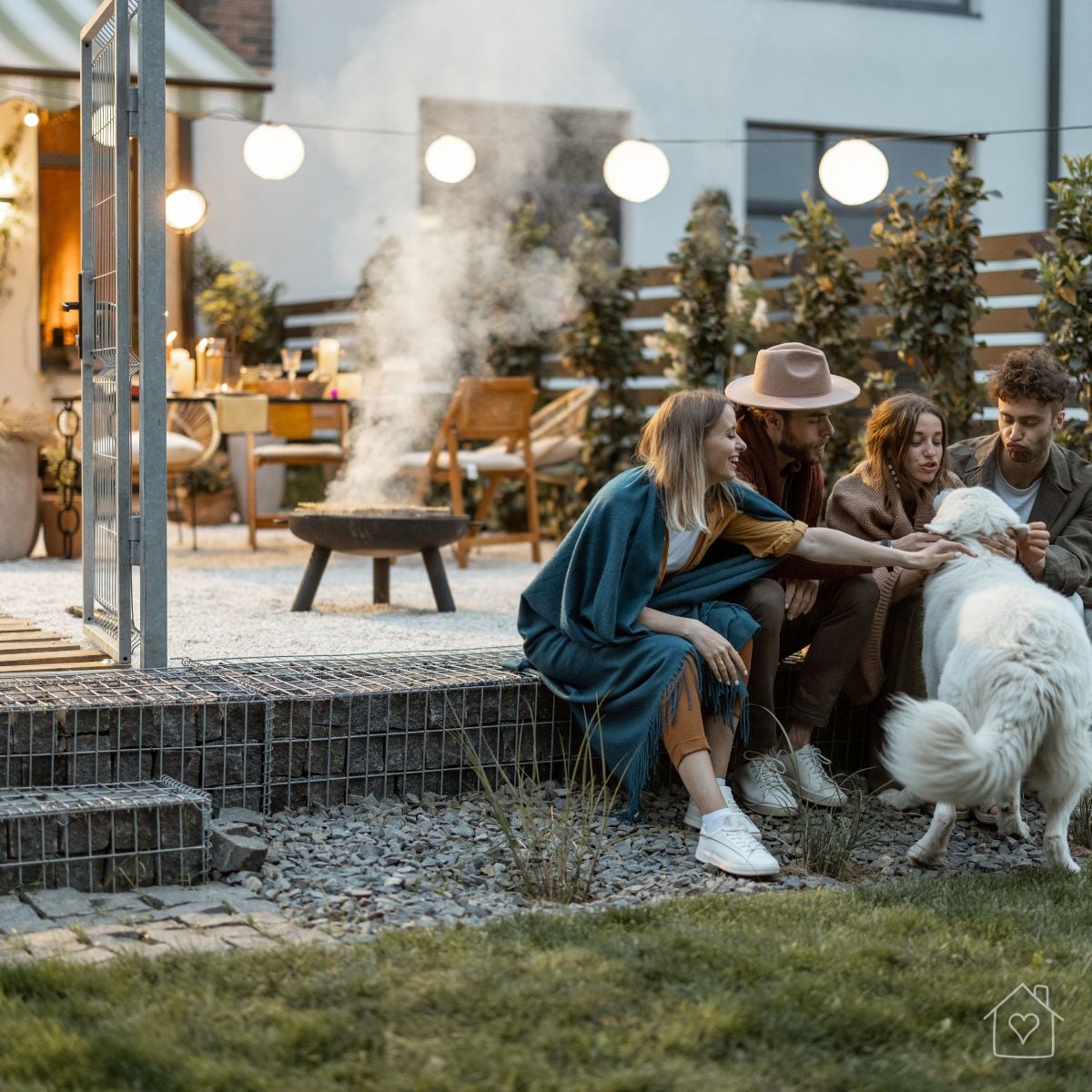 A group of friends and their dog relax together on a patio in the evening, with string lights and a fire pit glowing nearby