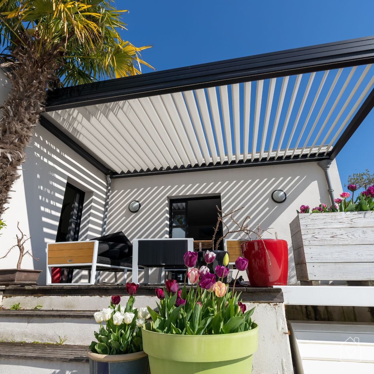 A black louvered pergola roof with the slats tilted open against a bright blue sky, mounted above a patio with potted tulips