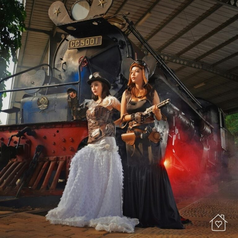 Two women in full steampunk costumes with steampunk goggles, top hat, and leather accessories standing in front of a vintage steam train