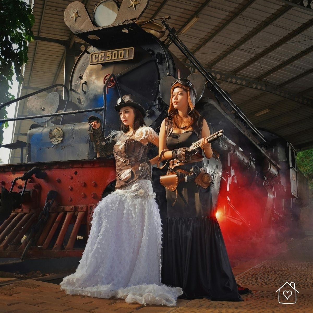 Two women in full steampunk costumes with steampunk goggles, top hat, and leather accessories standing in front of a vintage steam train