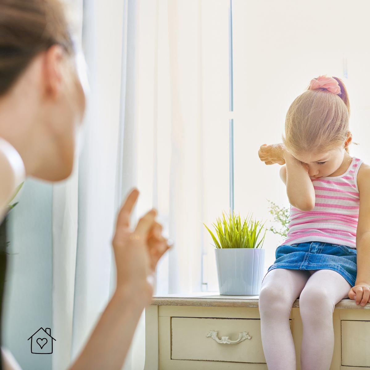 A mother points her finger while scolding her young daughter, who wipes her eyes and looks down, upset