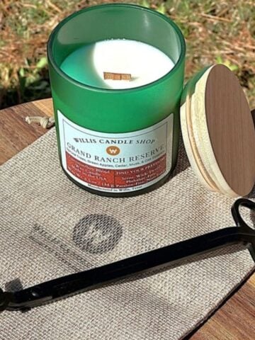 Green glass Willis Candle Shop candle labeled Grand Ranch Reserve, a coconut soy blend with spruce trees, green apples, cedar, musk, and oak essence, shown with a wood wick and trimming scissors