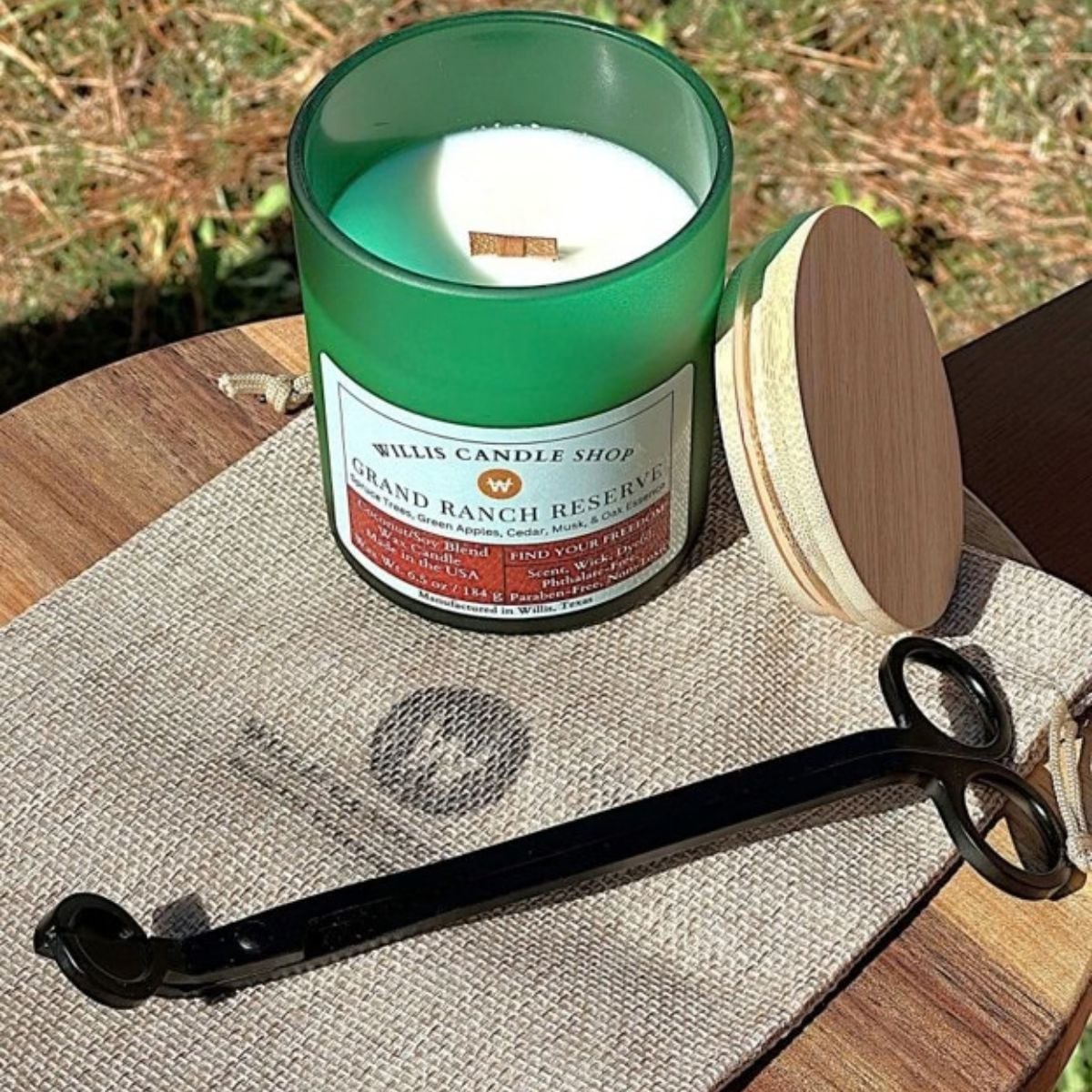 Green glass Willis Candle Shop candle labeled Grand Ranch Reserve, a coconut soy blend with spruce trees, green apples, cedar, musk, and oak essence, shown with a wood wick and trimming scissors