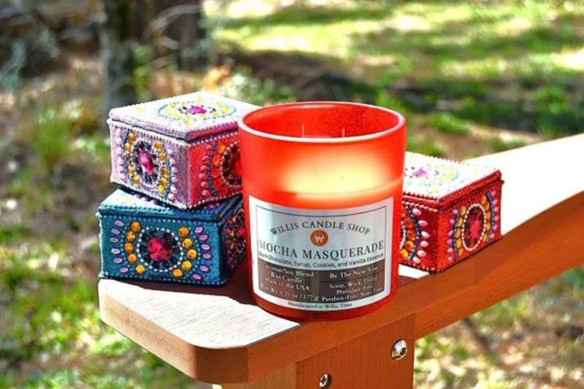 Orange glass Willis Candle Shop candle labeled Mocha Masquerade, a coconut soy blend with dark chocolate, syrup, cookies, and vanilla essence, styled next to two colorful jeweled trinket boxes