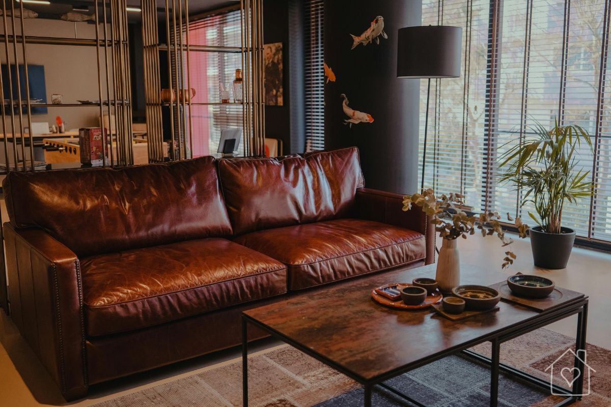 Deep oxblood leather sofa in a dark, modern living room with large windows and a rustic wood coffee table