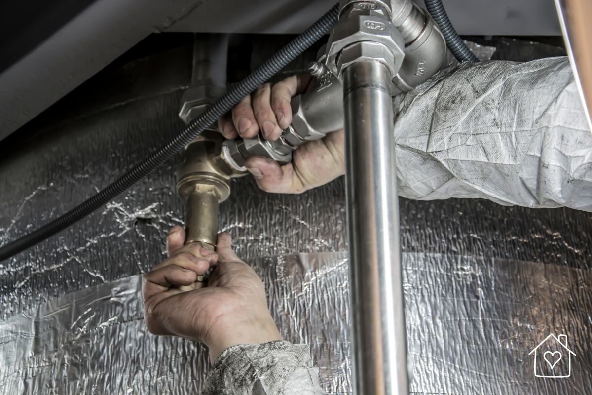 Two hands tightening a brass water valve fitting on a metal pipe beside foil insulation in a tight space