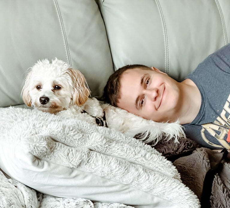 Small white dog with tan ears cuddling beside a smiling family member on the couch under a cozy blanket.
