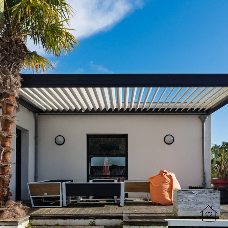 Louvered pergola with a black frame and angled white louvers over patio seating beside a white house and palm tree