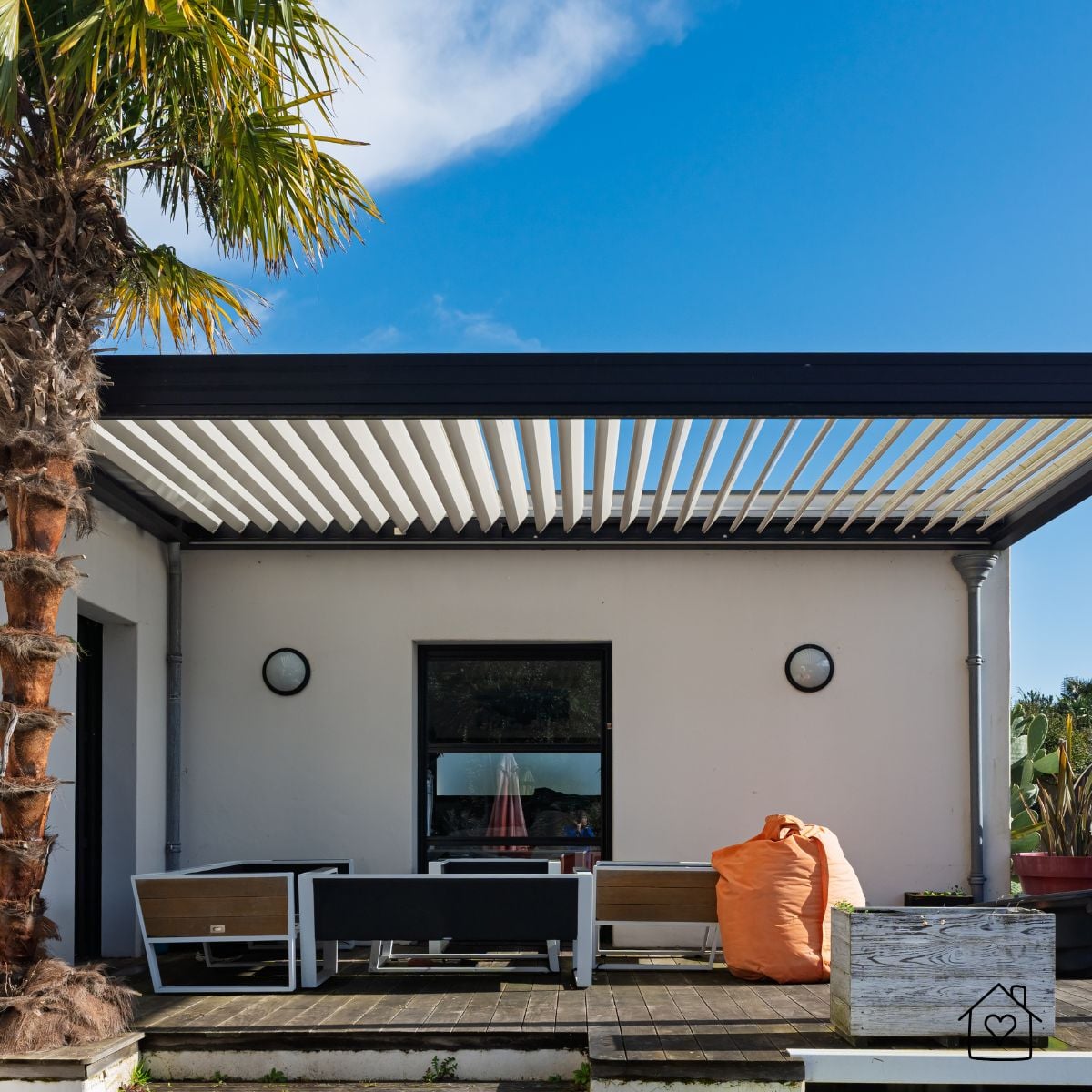 Louvered pergola with a black frame and angled white louvers over patio seating beside a white house and palm tree
