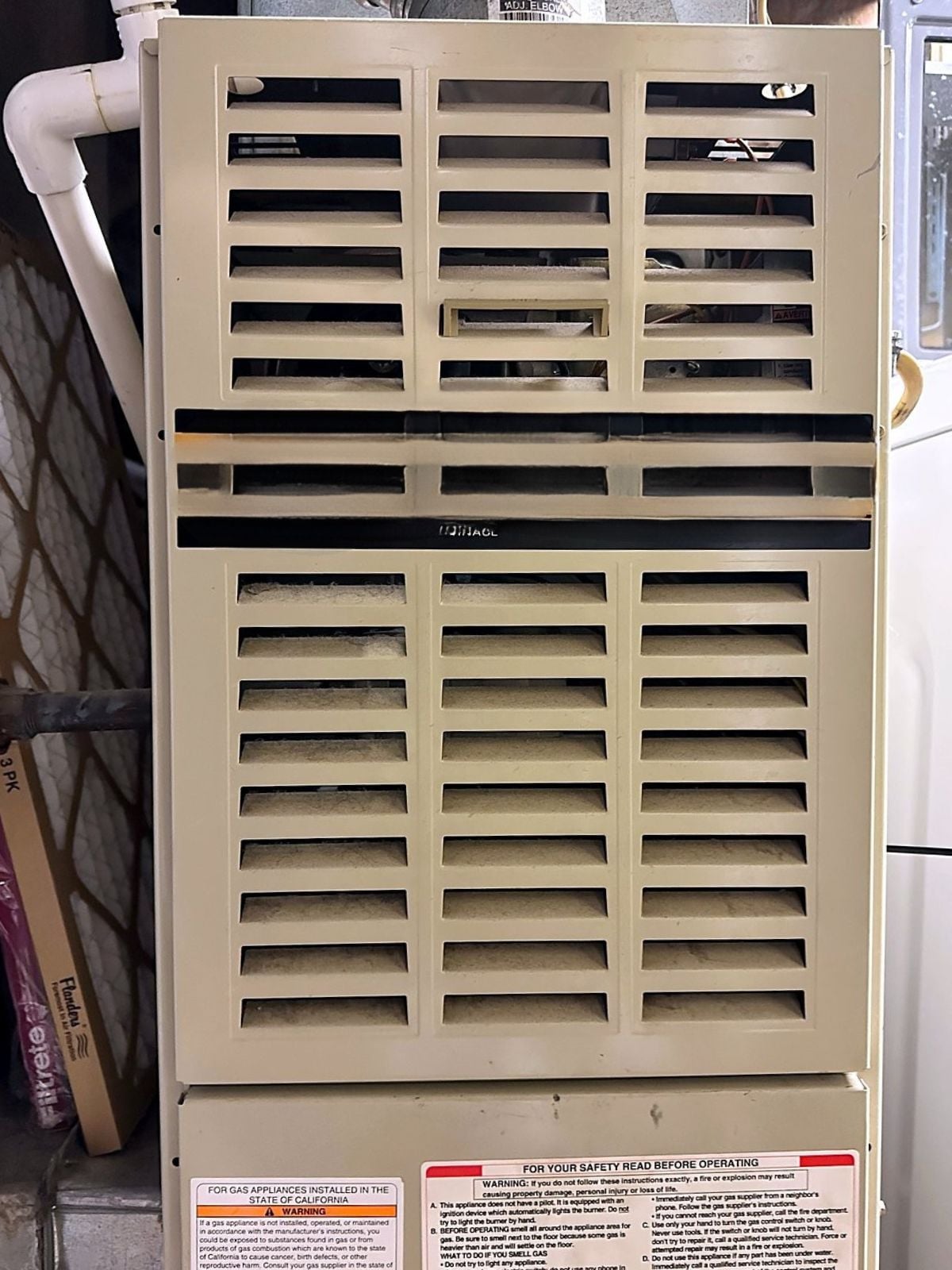 Front panel of a home gas furnace with dust on the louvered vents, and a pack of replacement air filters leaning beside it