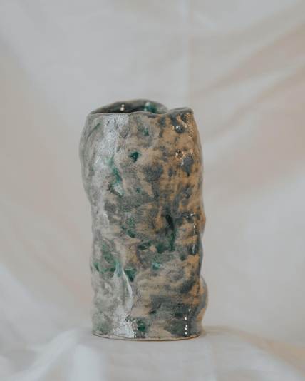 Textured ceramic vase with a speckled green and gray glaze on a white fabric backdrop