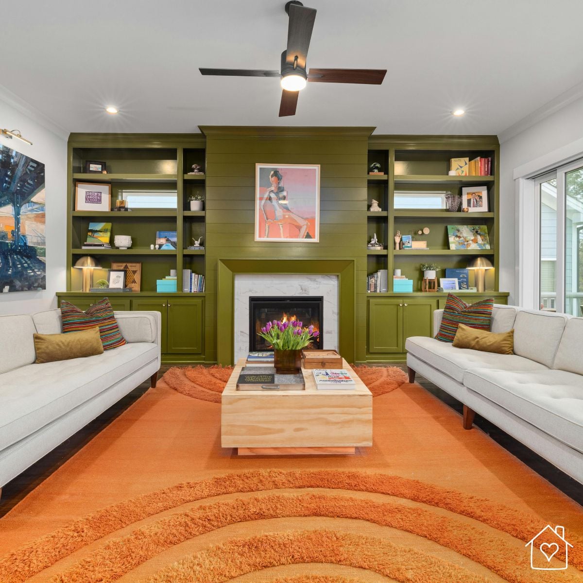 Living room with a lit fireplace centered between olive green built-in shelves, two cream sofas facing each other, brass lamps on the shelves, and an orange rug