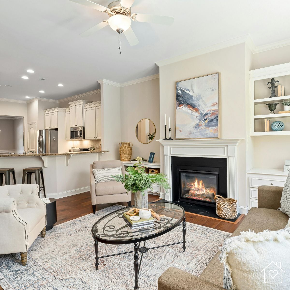 Neutral living room with a glowing fireplace, woven baskets, a fringed throw pillow, a soft patterned rug, and fresh greenery on a glass coffee table